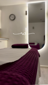Spa treatment bed with white sheet and purple textured blanket in a softly lit room; mirror on the wall reflects part of the space.